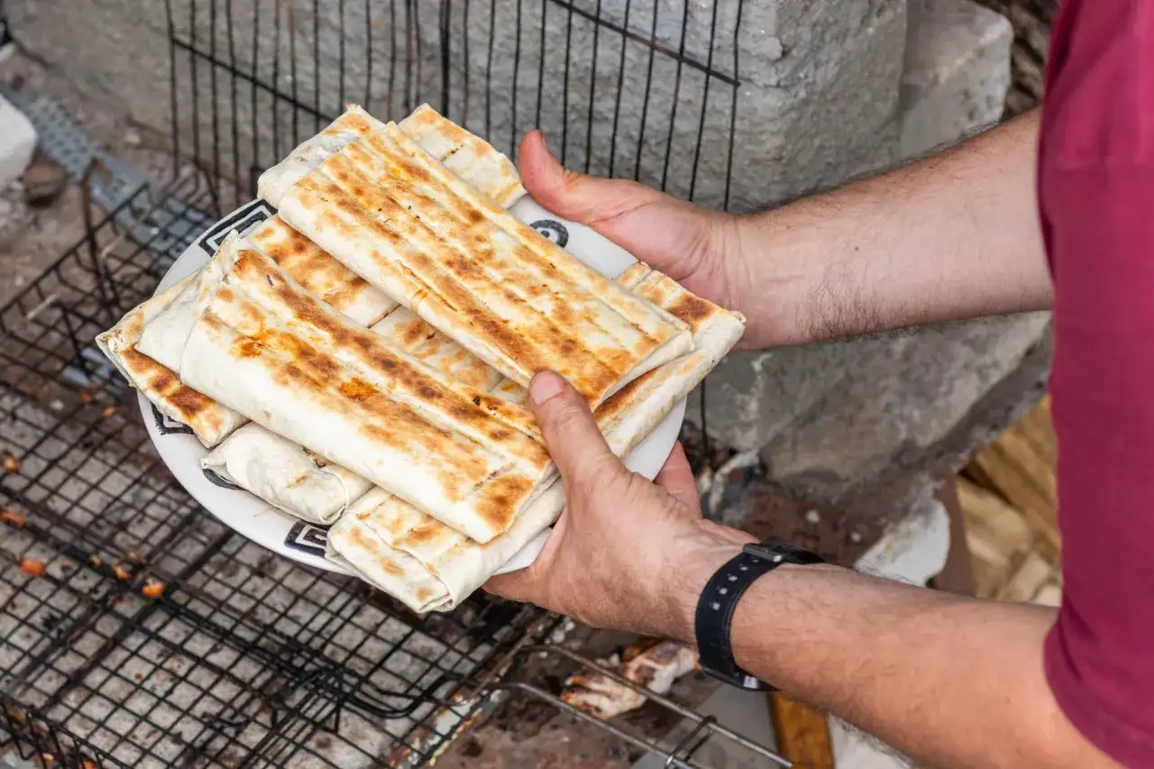 Talerz z grillowanymi wrapami kebab, trzymany przez ręce na tle rusztu i kamieni.